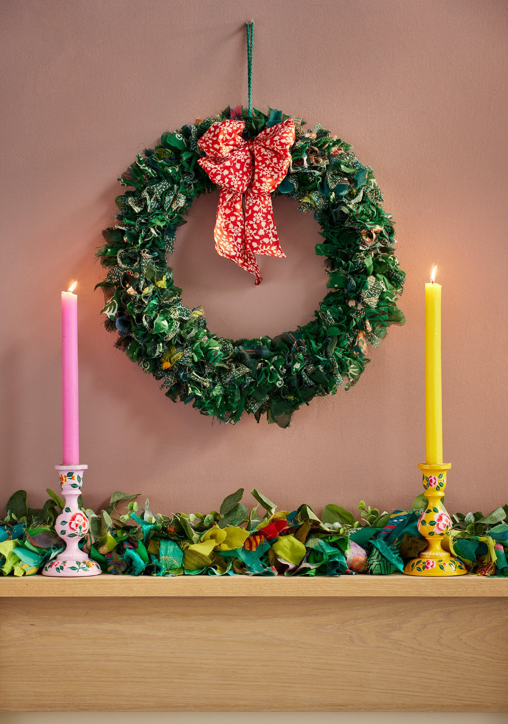 Decorative wreath with a red bow and candles on a mantelpiece against a pink wall.