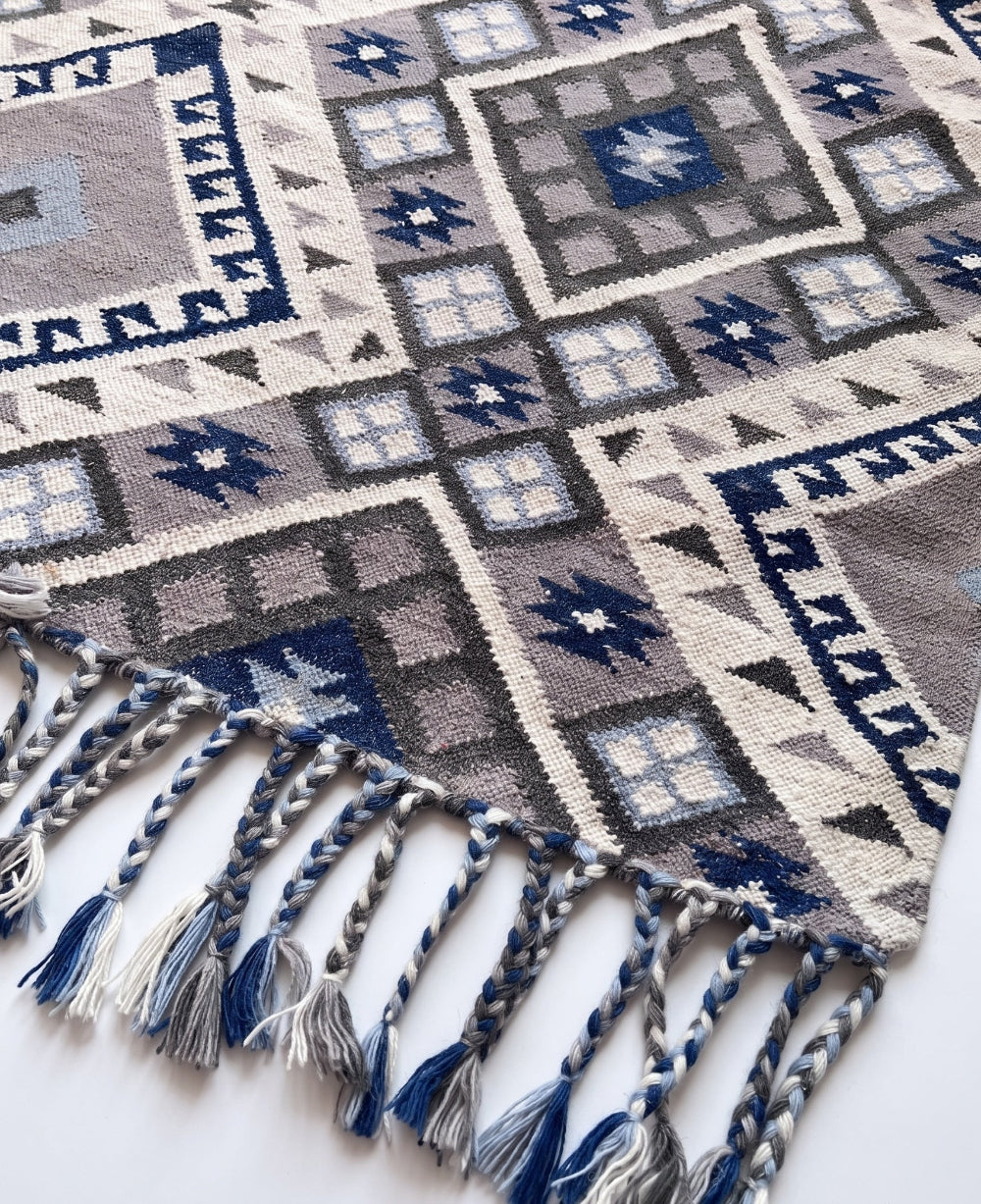 A close-up of a woven textile with geometric patterns in cream, grey, navy, and light blue, featuring braided fringe tassels along one edge.