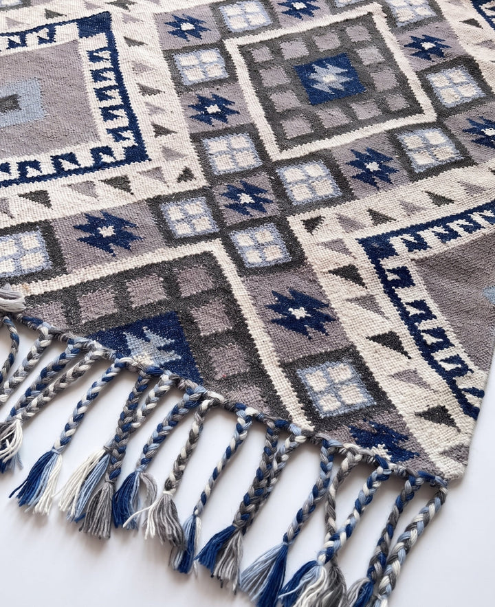A close-up of a woven textile with geometric patterns in cream, grey, navy, and light blue, featuring braided fringe tassels along one edge.