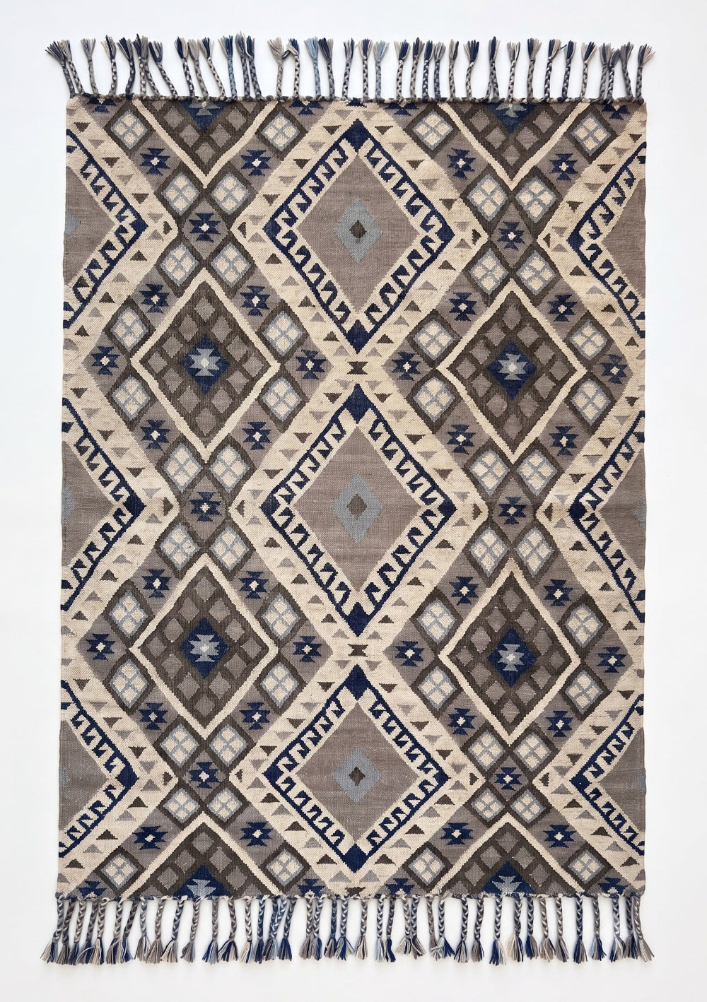 A rectangular woven rug with a geometric diamond pattern in beige, brown, grey, and navy blue, featuring dark blue fringe on the top and bottom edges.