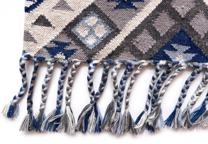 Close-up of a woven textile with braided fringe, featuring geometric diamond and triangle patterns in blue, grey, beige, and white on a plain white background.