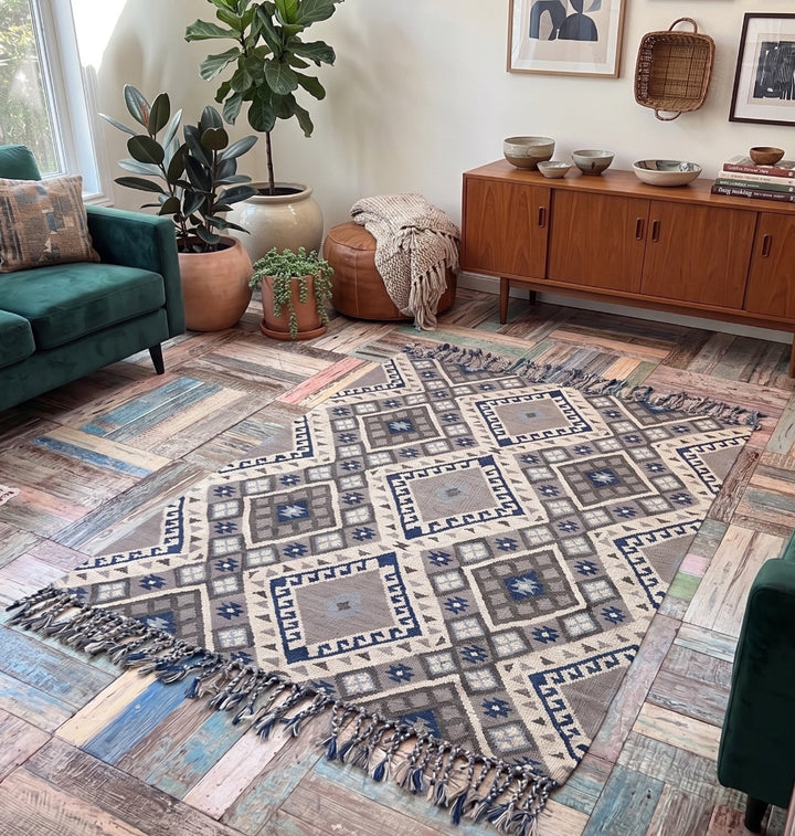 A rectangular area rug with a distressed geometric tribal pattern in blue, beige, and grey, styled on a rustic wood plank floor in a cosy living room setting.