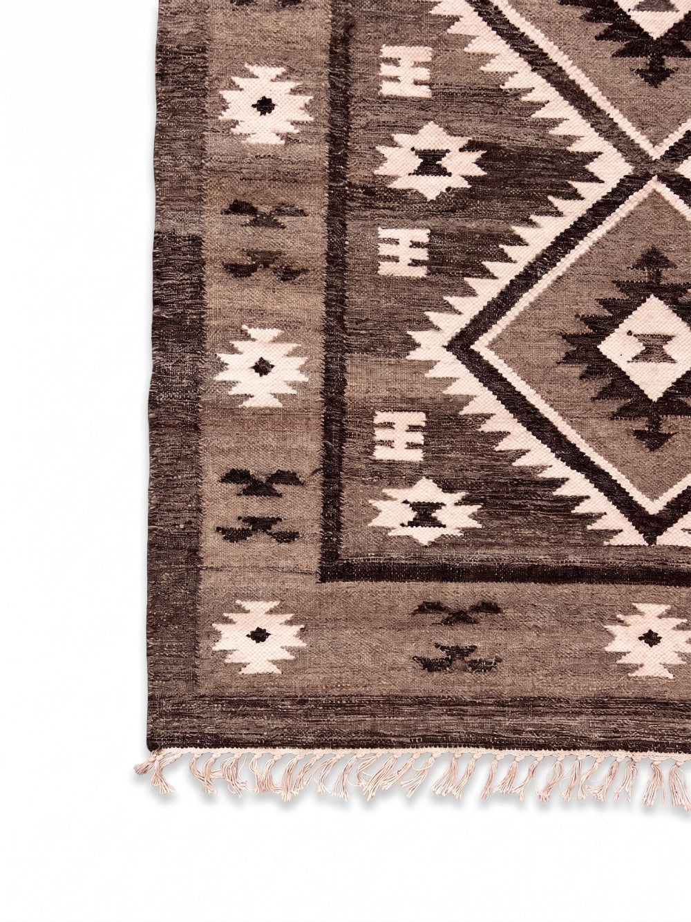 A close-up of a woven rug corner showing a geometric tribal pattern in brown, beige, and grey, with a cream-coloured fringe at the bottom.