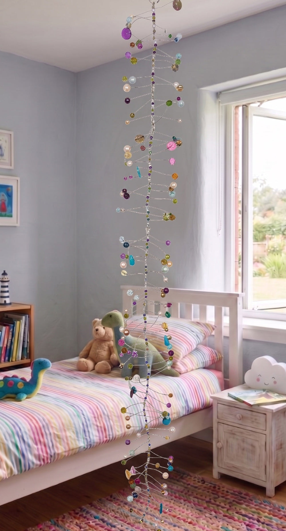 A colourful hanging mobile with small round beads and metallic accents hangs in front of a child's bed in a bedroom. The room has light grey walls, a white bed frame, striped pastel bedding, a teddy bear, and a window on the right.