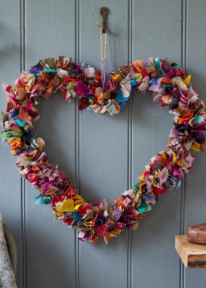 A heart-shaped fabric wreath made from colourful, textured cloth strips hangs on a blue-grey panelled wall. The scene includes a small vase with dried stems on the left, a window on the right, and a wooden shelf with books and glasses in the foreground.