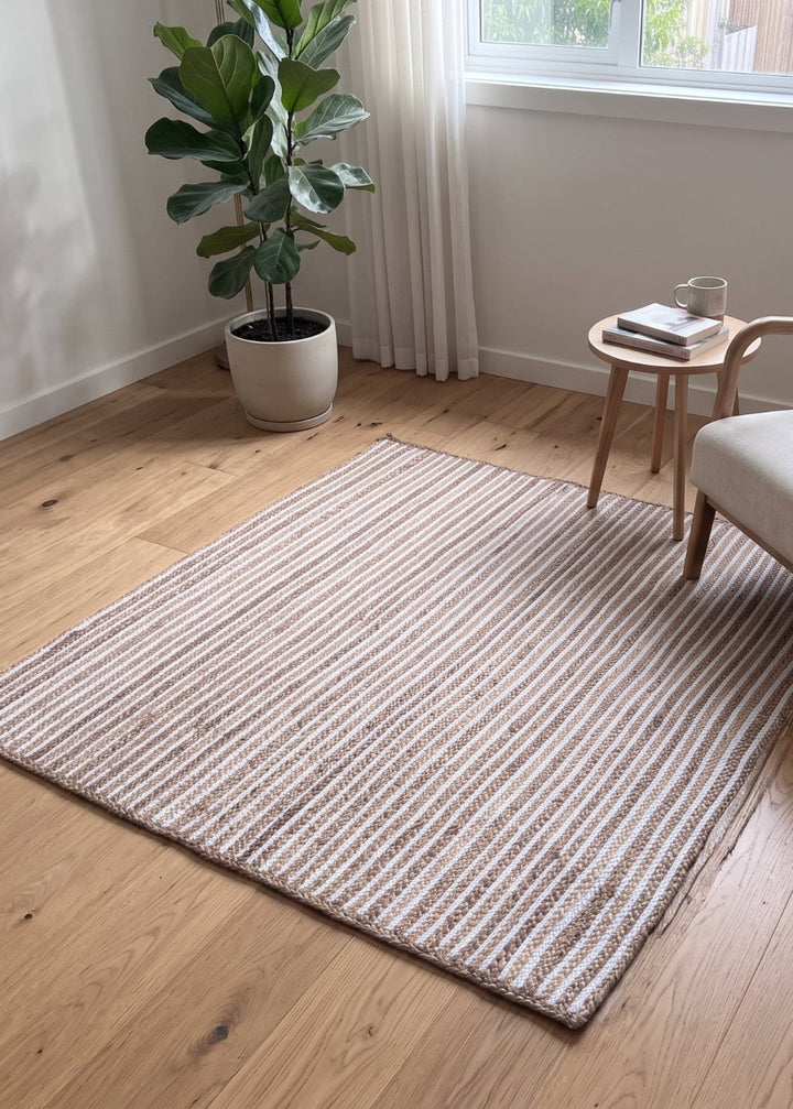 A rectangular striped area rug in beige and brown tones, styled in a bright, modern room with a light wood floor, a wooden armchair, a small side table, and a potted plant.