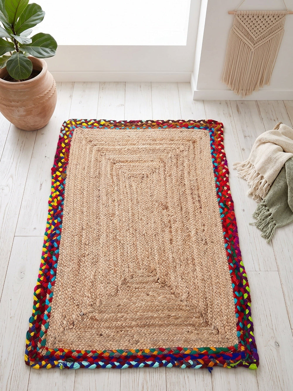 Beige braided rug with colourful border on a light wooden floor, next to a plant and a woven basket.