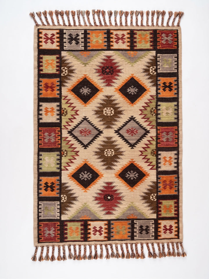 A rectangular woven area rug with a Southwestern geometric pattern, featuring a cream beige centre, a dark brown border, and orange, red, and gray accents, finished with fringed tassels on the top and bottom.
