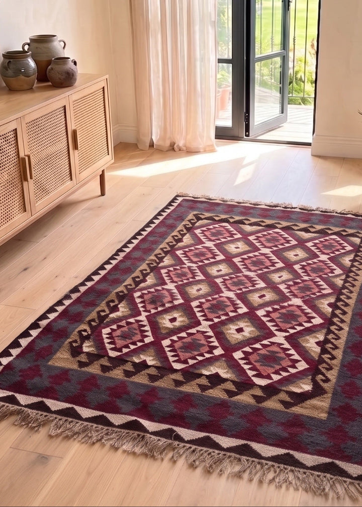 A rectangular area rug with a geometric tribal pattern in red, beige, and blue, styled on a light wood floor in a bright, modern room with a wooden sideboard and an open door.