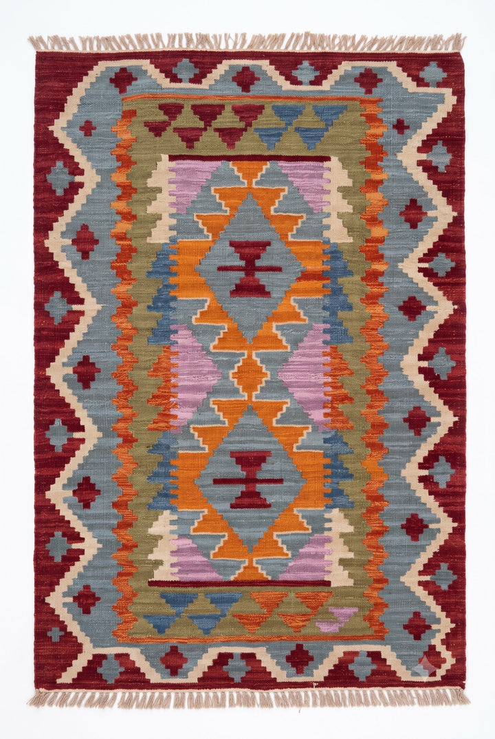A rectangular woven rug with a Southwestern geometric pattern, featuring a central diamond motif in orange, yellow, and purple, surrounded by blue, green, and red bands, finished with fringe on the top and bottom edges.