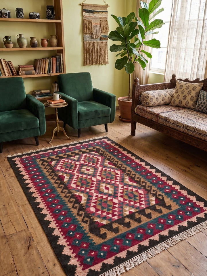 A rectangular area rug with a geometric tribal pattern in pink, beige, black, and teal, styled on a wooden floor in a cosy living room with green armchairs and a wooden sofa.