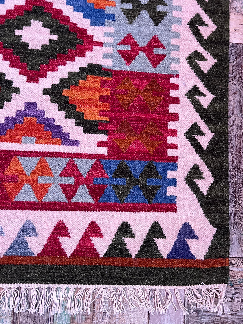 A close-up of a woven geometric rug corner showing a multicolor pattern with pink, red, blue, orange, black, and white sections, finished with white fringe along the bottom edge.