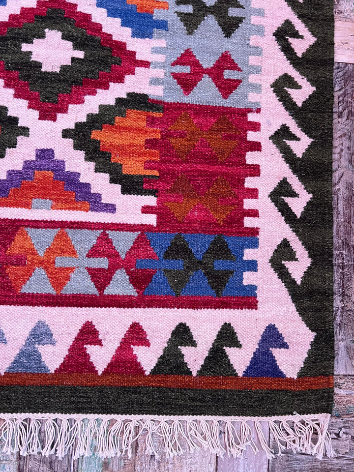 A close-up of a woven geometric rug corner showing a multicolor pattern with pink, red, blue, orange, black, and white sections, finished with white fringe along the bottom edge.