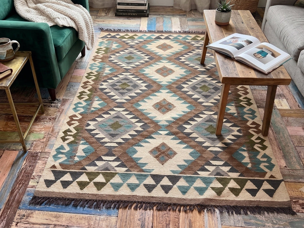 A rectangular area rug with a geometric diamond pattern in beige, brown, teal, and blue, styled in a cozy living room next to a green armchair and a gray sofa.