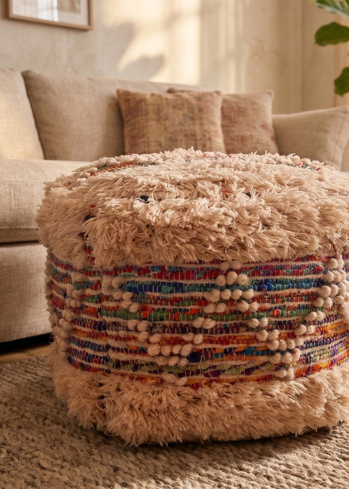A round pouf ottoman with a shaggy beige faux fur top and a colourful braided rug base, styled in a warm, modern living room next to a beige sofa.