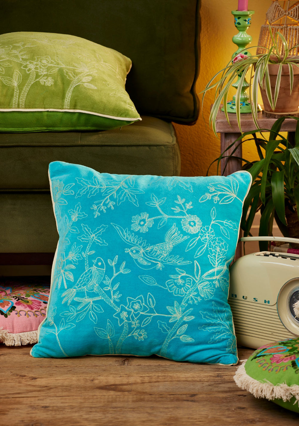 Turquoise decorative pillow with floral pattern on a wooden surface, surrounded by green and pink pillows.