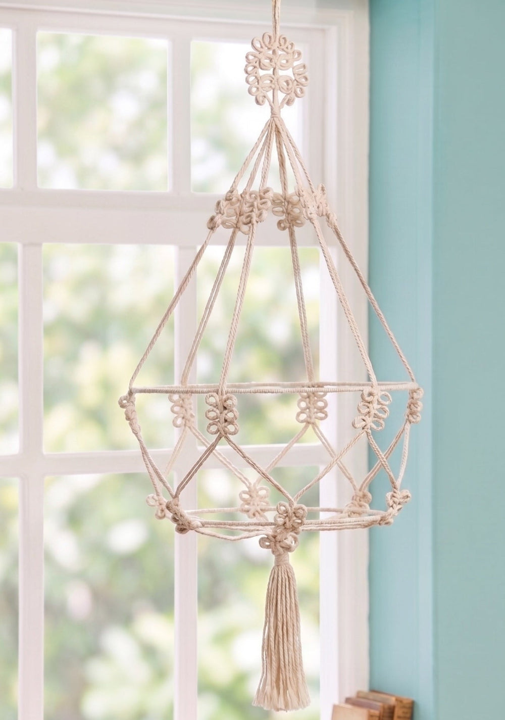 Decorative macrame hanging with tassels against a light blue wall and window.