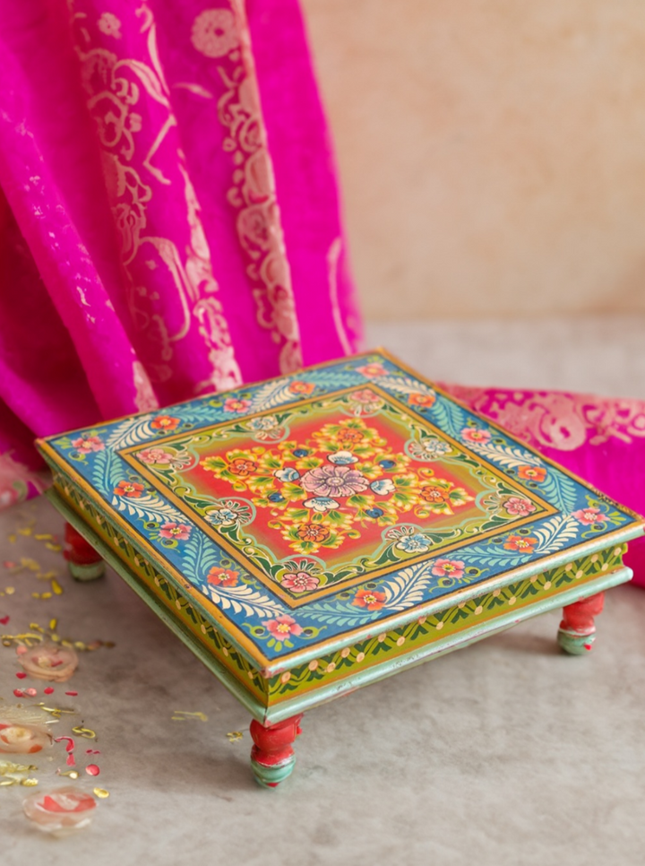 A handcrafted mango wood bajot table with a colourful, floral hand-painted design on the tabletop, supported by four curved legs with red and green accents.