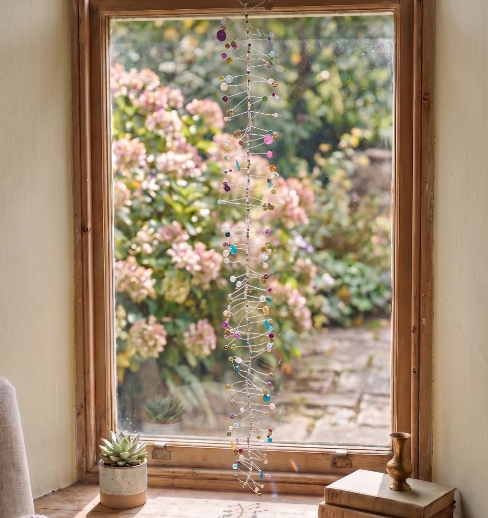 A long, vertical hanging string ornament with many small colourful beads and metallic links is displayed in front of a wooden window. Outside the window, pink flowers and green foliage are visible, and small potted plants sit on the windowsill.