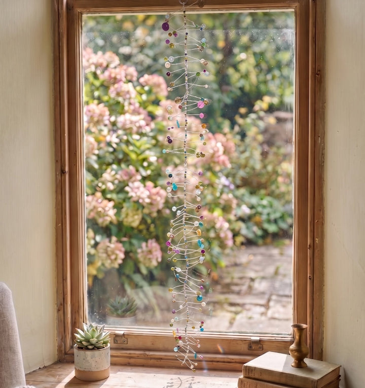 A long, vertical hanging string ornament with many small colourful beads and metallic links is displayed in front of a wooden window. Outside the window, pink flowers and green foliage are visible, and small potted plants sit on the windowsill.