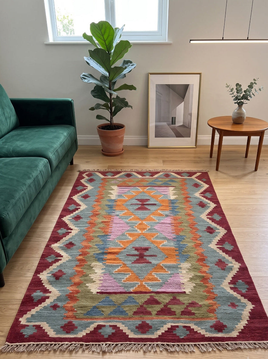 A colourful geometric area rug with fringe, displayed on a light wood floor in a modern living room setting next to a green sofa.