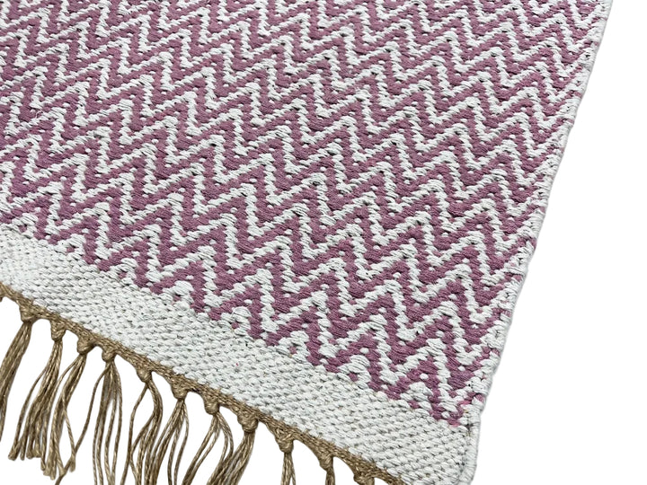 A close-up of a woven textile with a repeating chevron pattern in dusty mauve and off-white, finished with a tan fringed edge on a white background.