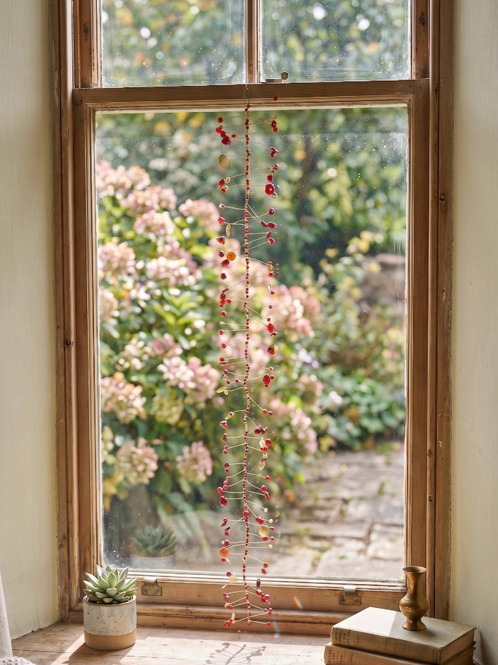 A long, delicate red berry garland hangs vertically in front of a rustic wooden window, with a garden of pink flowers visible outside. The window sill features a small potted succulent and a stack of vintage books.