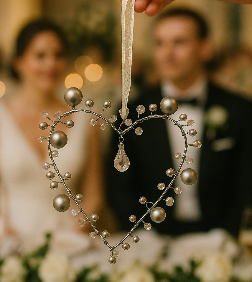 Decorative heart-shaped ornament with pearls and a teardrop pendant, held by a hand with a blurred wedding couple in the background.
