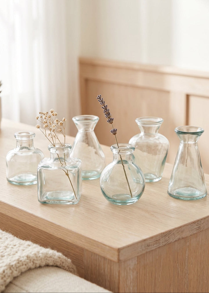 Six small clear glass vases in different shapes arranged on a light wood table in a softly lit room.