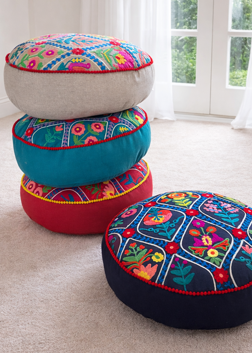 Stack of colourful embroidered small cushions in a room with white curtains and greenery outside.