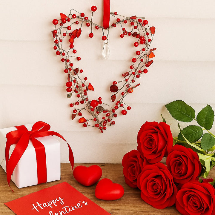 Heart-shaped wreath with red berry beads, gift box, roses, and 'Happy Valentine's' card on a wooden surface.