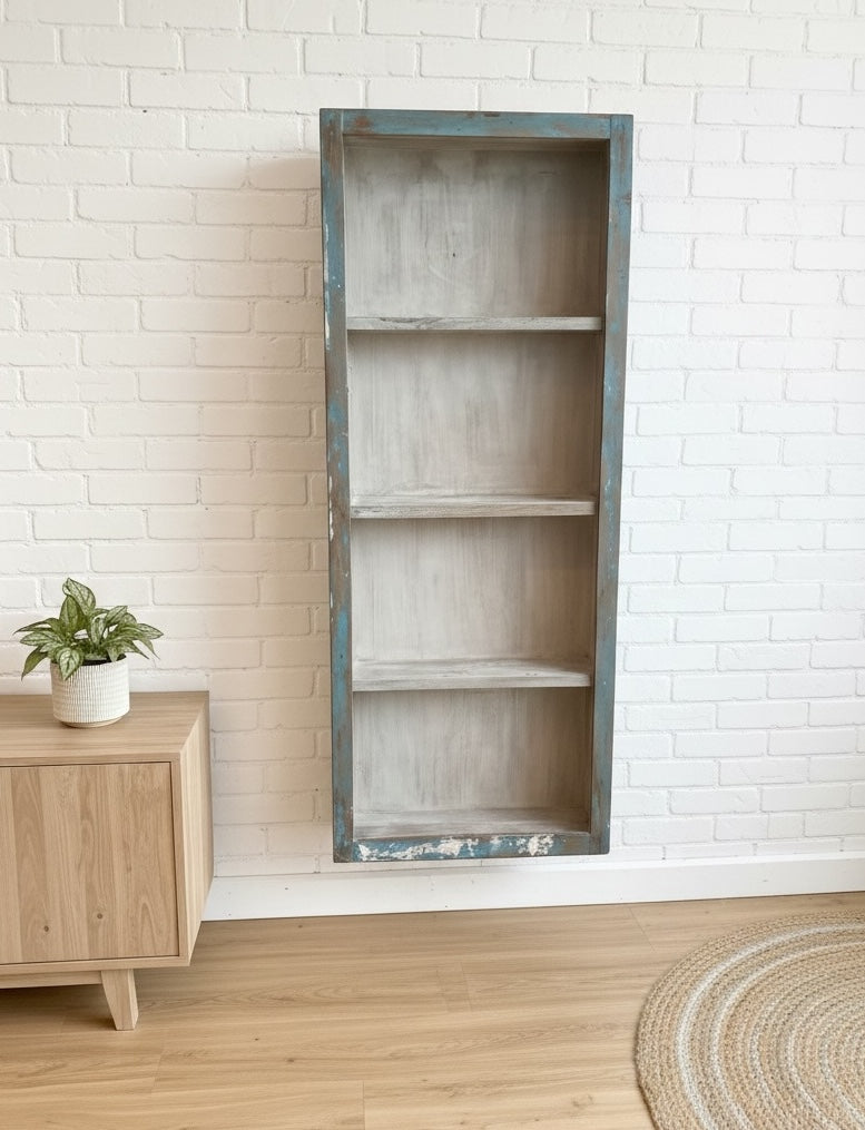 Tall narrow bookshelf display cabinet against a white brick wall with a wooden cabinet and plant in the foreground.