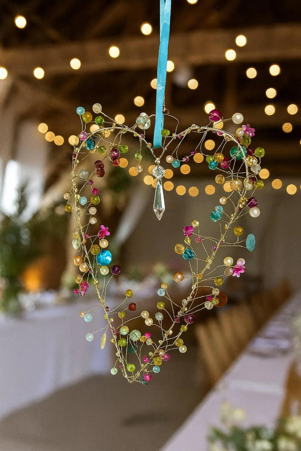 Decorative heart-shaped hanging with colourful beads and a blue ribbon against a blurred indoor background with lights.