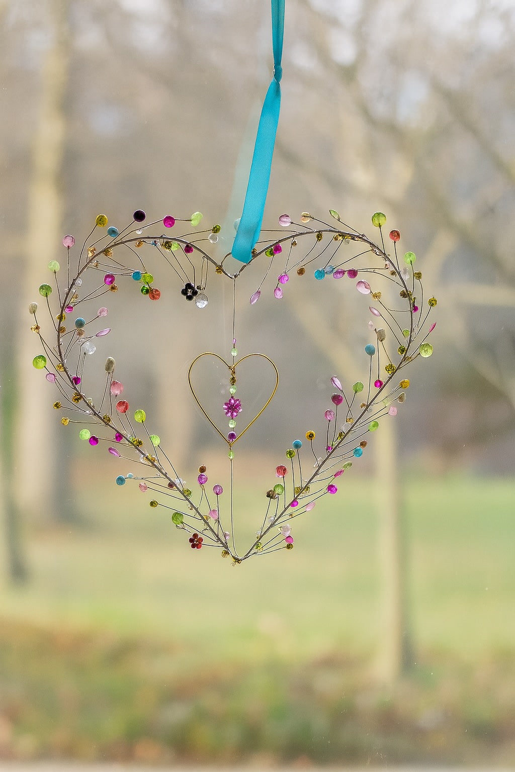 Decorative heart-shaped ornament with colourful beads and a blue ribbon against a blurred natural background.