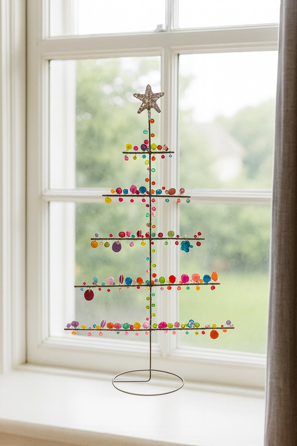 Decorative window art resembling a Christmas tree with colourful beads and a star on top, placed in front of a window.