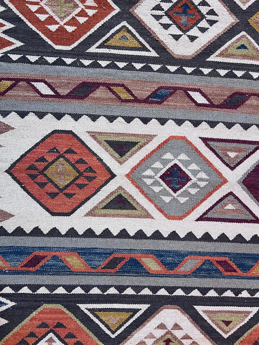 A close-up of a woven geometric rug with Southwestern-style patterns in rust red, navy, gray, cream, and olive green.