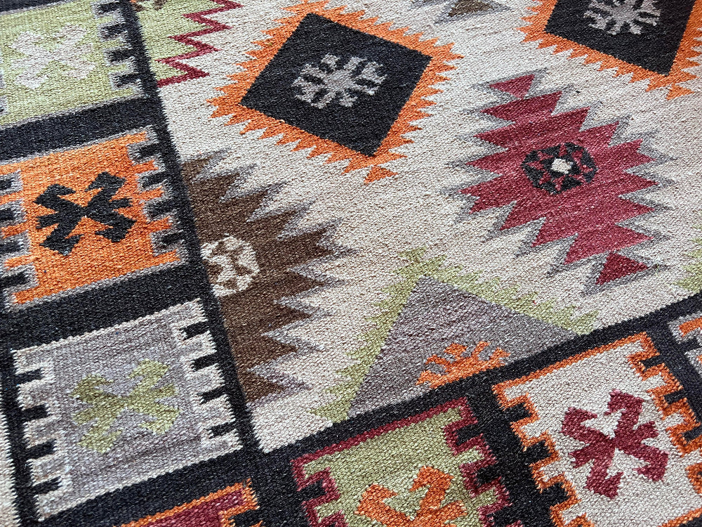 Close-up of a woven rug with a Southwestern geometric pattern in black, orange, red, gray, brown, and sage green on a cream background.