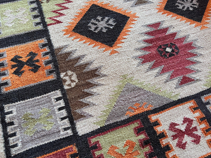 Close-up of a woven rug with a Southwestern geometric pattern in black, orange, red, gray, brown, and sage green on a cream background.