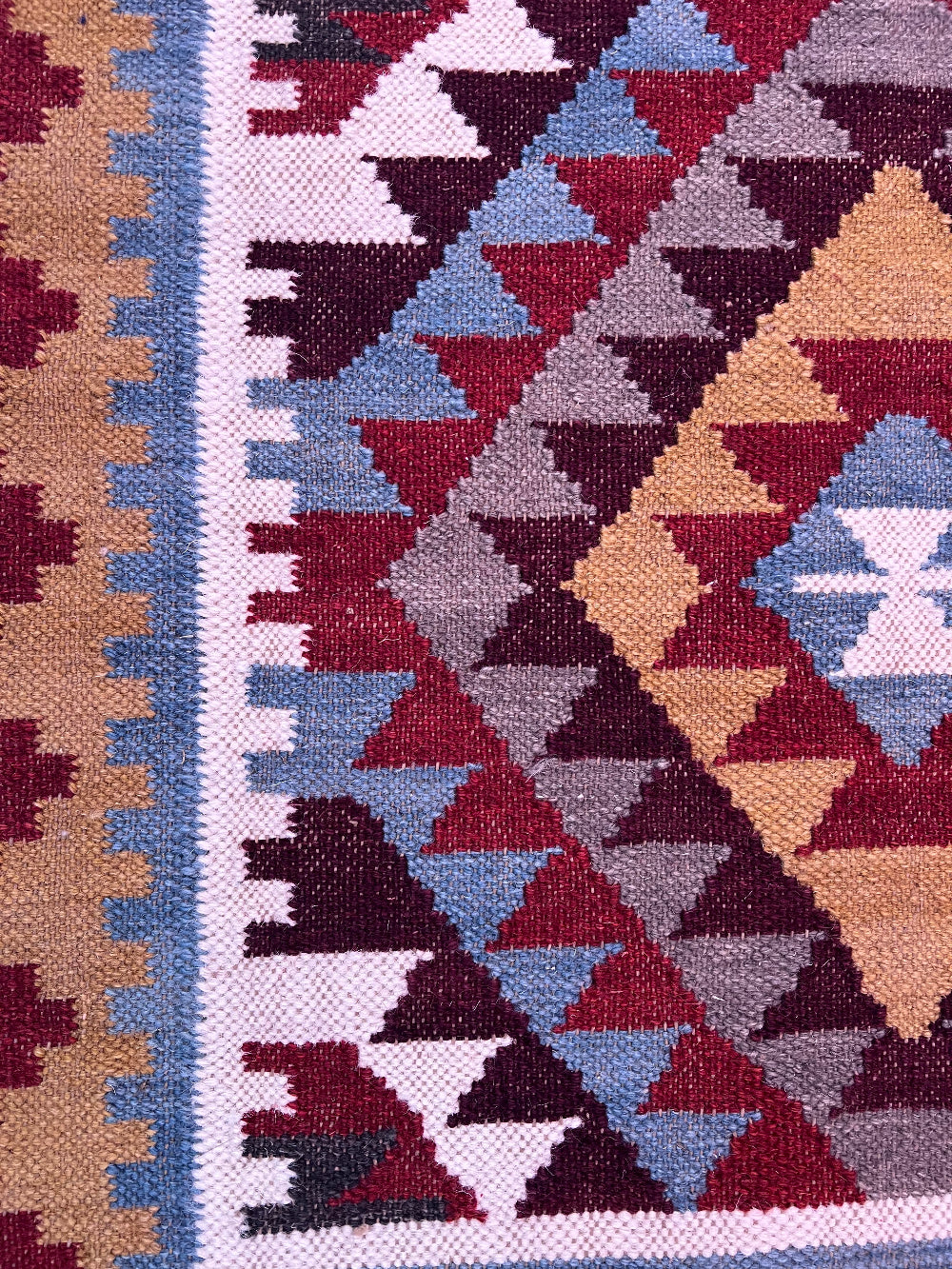 Close-up of a woven geometric textile with repeating diamond and triangle motifs in rust red, beige, light blue, white, and dark plum.
