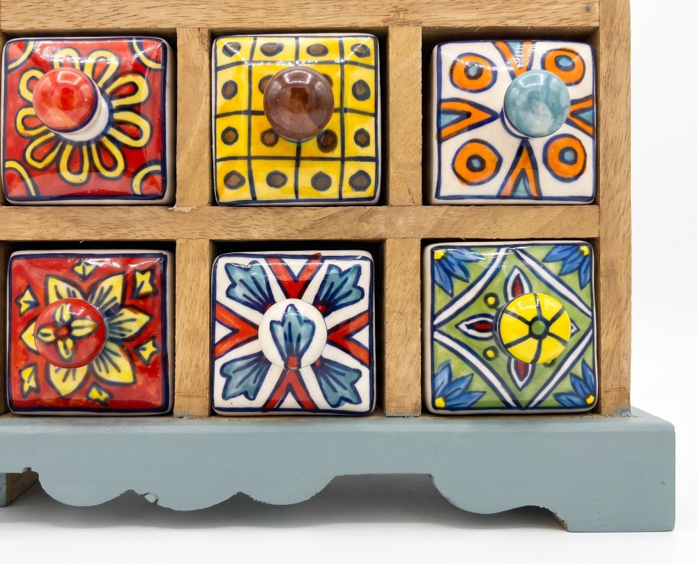 A wooden spice rack with six square ceramic drawer fronts decorated in colorful hand-painted patterns, set against a plain white background.