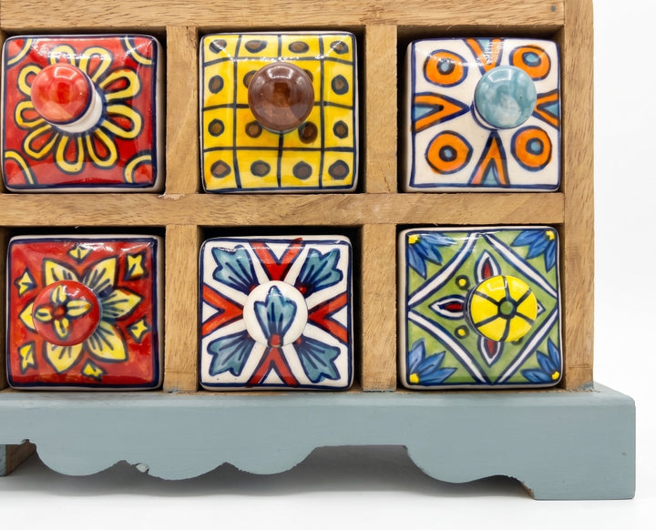 A wooden spice rack with six square ceramic drawer fronts decorated in colorful hand-painted patterns, set against a plain white background.
