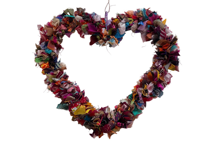 A heart-shaped fabric wreath made from colourful shredded paper or ribbon, hanging on a white tabletop with a small paper tag at the top.