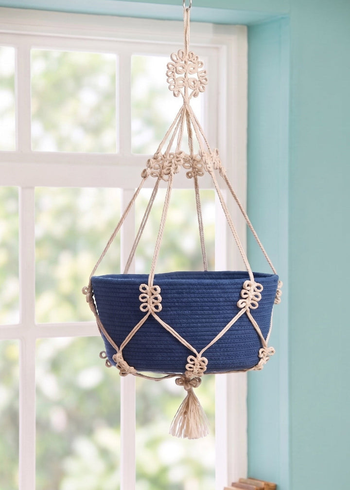 Blue woven basket with gold accents hanging from a window on a macrame jute plant holder