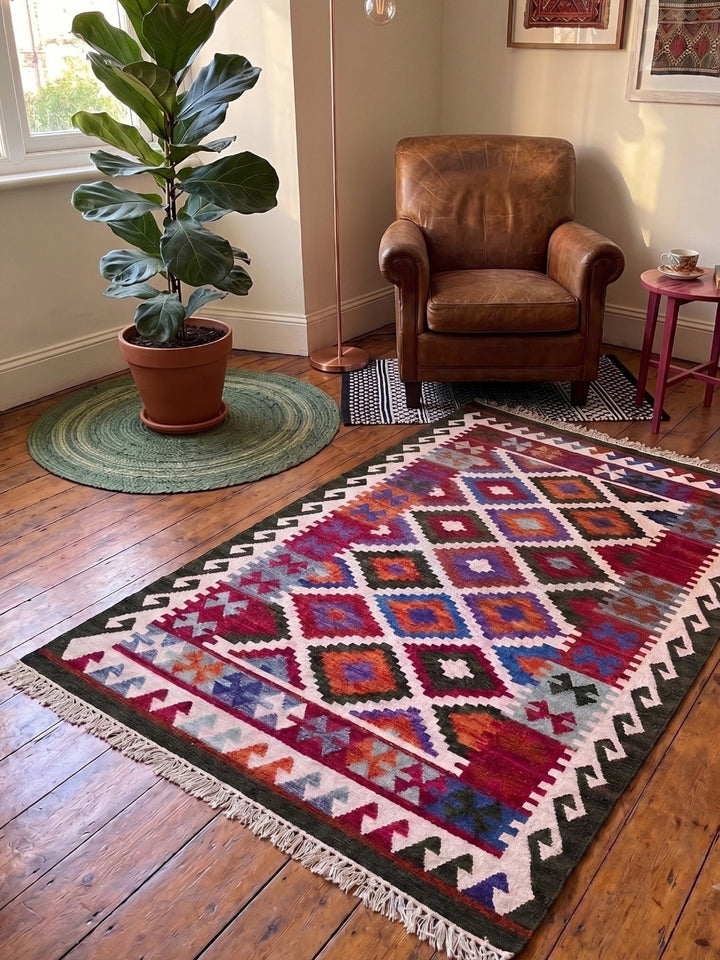 A rectangular kilim area rug with a colorful geometric diamond pattern and white fringe, styled in a warm, rustic living room next to a brown leather armchair, a potted fiddle-leaf fig plant, and a wooden floor.