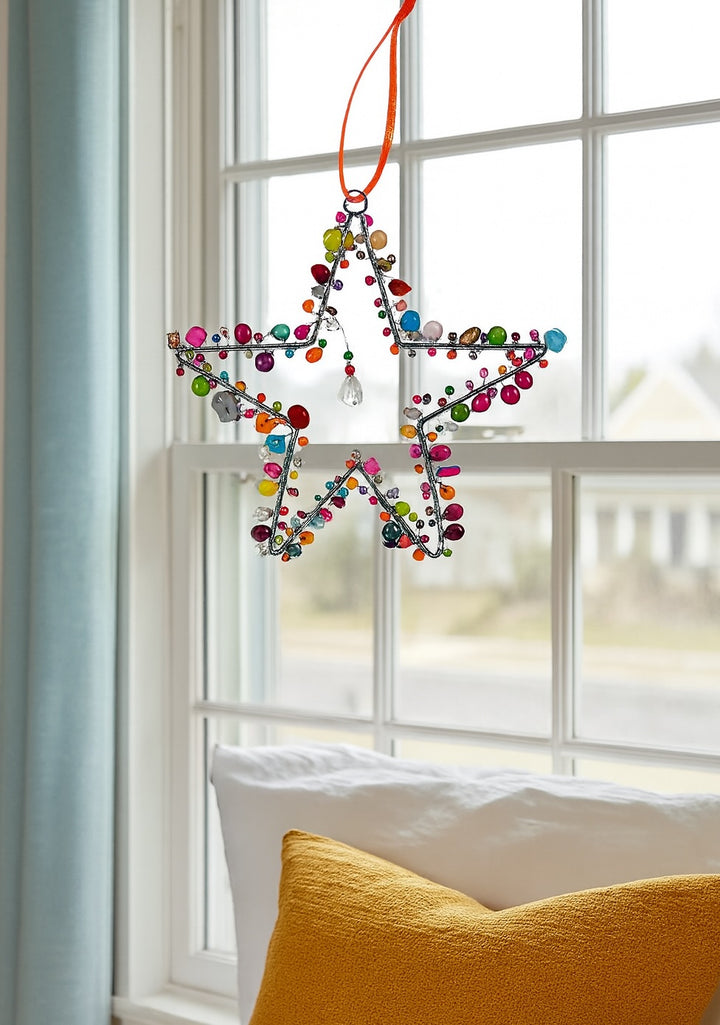 Colourful star-shaped decoration hanging by a window with a yellow pillow on a chair.